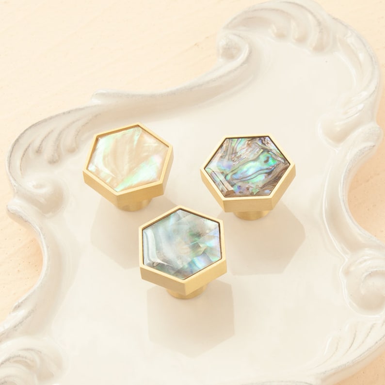 May include: Three hexagonal cabinet knobs with a gold finish and iridescent mother-of-pearl inlays. The knobs are arranged in a triangular pattern on a white surface.