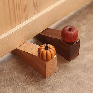 Pode incluir: Dois batentes de porta de madeira, um claro e outro escuro, cada um encimado por uma pequena fruta decorativa. O batente claro tem uma pequena abóbora laranja, e o batente escuro tem uma maçã vermelha. Os batentes de porta estão sobre uma superfície de cor neutra.