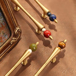 May include: Close-up of several gold-toned cabinet pulls with decorative gemstone accents. Each pull features a different gemstone, including blue, red, green, and tiger's eye, and is mounted on a brown surface.