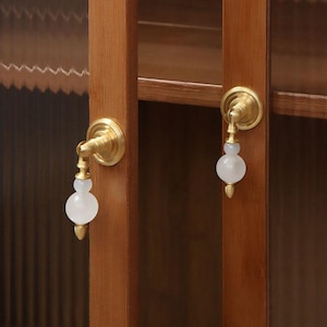 May include: Close-up of cabinet door hardware. The hardware features a brass finish with a white, spherical, and elongated handle. The cabinet doors are made of wood and have a light brown color.