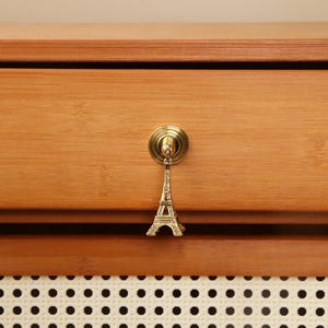 Peut inclure: Un tiroir en bois avec un bouton en laiton orn&eacute; d'un charm miniature de la Tour Eiffel. Le tiroir est en bois brun clair et poss&egrave;de un panneau d&eacute;coratif blanc et noir en bas.