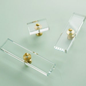 May include: Three clear glass cabinet knobs with gold metal accents. The knobs are rectangular and have a simple design.