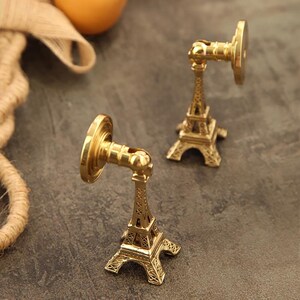 Peut inclure: Deux crochets muraux d&eacute;coratifs dor&eacute;s en forme de Tour Eiffel. Les crochets ont une base circulaire et un bras pivotant. La partie de la Tour Eiffel est finement d&eacute;taill&eacute;e. Les crochets sont sur une surface gris fonc&eacute;.