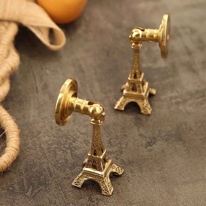 Peut inclure: Deux crochets muraux d&eacute;coratifs dor&eacute;s en forme de Tour Eiffel. Les crochets ont une base circulaire et un bras pivotant. La partie de la Tour Eiffel est finement d&eacute;taill&eacute;e. Les crochets sont sur une surface gris fonc&eacute;.