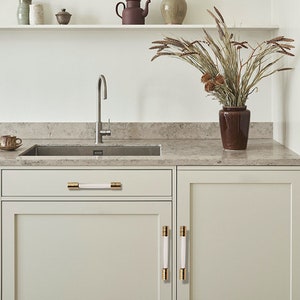 以下が含まれることがあります： 金と白のキャビネットノブが付いた白いキッチンのキャビネット。キャビネットは、ステンレス製のシンクと蛇口のあるモダンなキッチンにあります。