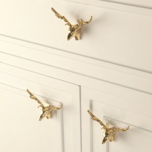 Op de afbeelding: Drie gouden kastgrepen in de vorm van een hertenkop met geweien. De grepen zijn gemonteerd op een witte kast.