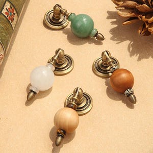 May include: A selection of cabinet knobs with brass fittings. The knobs have spherical designs in green jade, white stone, and wood. The brass hardware has an antique finish, ideal for furniture restoration or custom projects. The knobs are suitable for various furniture styles.