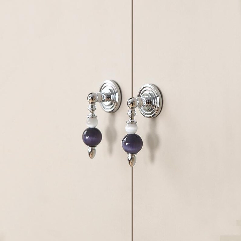 Pode incluir: Dois puxadores de arm&aacute;rio em metal prateado com uma placa de base circular e um design pendente. Cada puxador consiste numa pequena conta esf&eacute;rica branca encimada por uma conta esf&eacute;rica maior, roxo-escura, com efeito olho de gato.