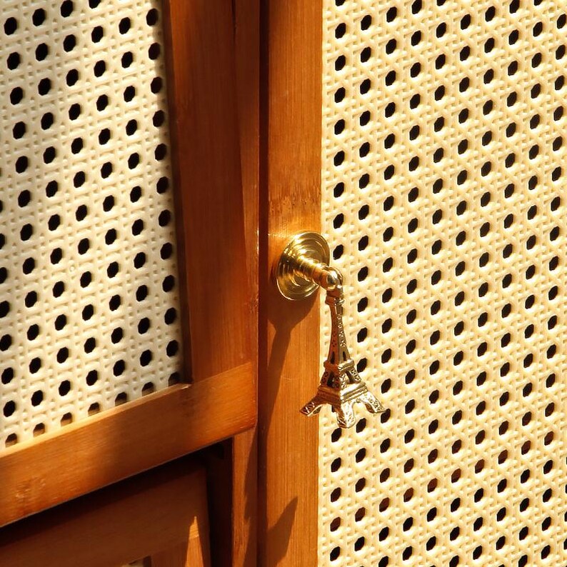 Peut inclure: Un charm dor&eacute; en forme de Tour Eiffel est suspendu &agrave; un bouton de meuble en laiton. Le bouton est mont&eacute; sur une porte d'armoire en bois avec un panneau tiss&eacute; de couleur cr&egrave;me. Le charm est une r&eacute;plique miniature du monument parisien embl&eacute;matique.