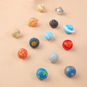 Könnte beinhalten: Eine Sammlung dekorativer Knöpfe in Planetenform. Jeder Knopf stellt einen anderen Planeten dar, mit unterschiedlichen Designs und Farben, darunter Blau, Orange, Gelb und Grau. Die Knöpfe sind auf einer hellrosa Oberfläche angeordnet.