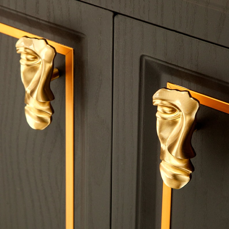 May include: Two gold-toned cabinet door pulls shaped like abstract faces with a textured surface. The pulls are mounted on a dark wood cabinet door with a gold trim.