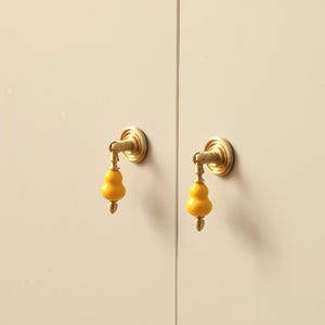 Peut inclure: Deux poignées de placard avec une finition en laiton et des poignées en céramique jaune vif en forme de gourde. Les poignées sont montées sur une porte d'armoire de couleur claire, offrant une touche de couleur et un élément de design unique.