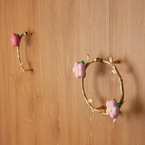 Può includere: Due maniglie decorative dorate. Una è una maniglia curva con un fiore rosa e foglie verdi. L'altra è una maniglia circolare con due fiori rosa e foglie verdi. Entrambe sono montate su una superficie di legno.