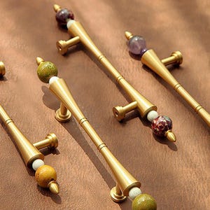 Op de afbeelding: Goudkleurige kastgrepen met decoratieve stenen accenten. Elke greep heeft een cilindrische handgreep met een uitlopende basis en een decoratieve stenen kraal aan de bovenkant. De stenen zijn in tinten groen, wit, paars en rood.