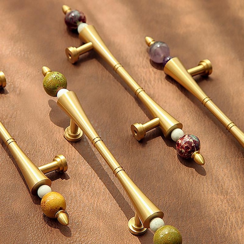 Op de afbeelding: Goudkleurige kastgrepen met decoratieve stenen accenten. Elke greep heeft een cilindrische handgreep met een uitlopende basis en een decoratieve stenen kraal aan de bovenkant. De stenen zijn in tinten groen, wit, paars en rood.