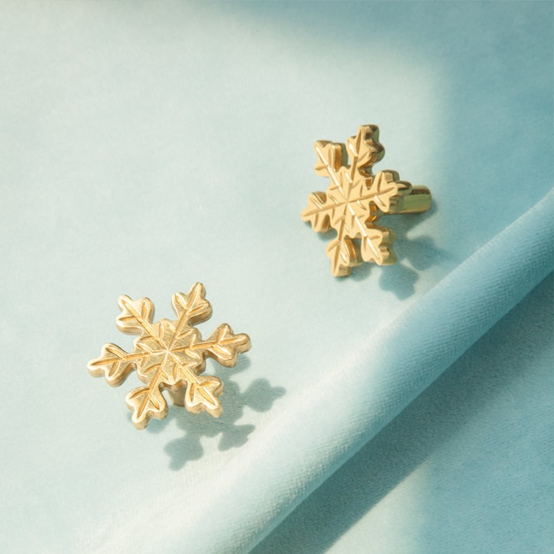 Op de afbeelding: Twee gouden trekknoppen in de vorm van sneeuwvlokken op een lichtblauwe achtergrond.