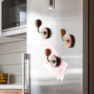 Puede incluir: Tres ganchos magnéticos de madera con un gancho de metal plateado y una perilla redonda de madera. Los ganchos están unidos a un refrigerador de acero inoxidable.