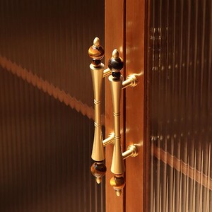 Op de afbeelding: Close-up van een kastdeur met twee sierlijke messing handgrepen. De handgrepen hebben een decoratief ontwerp met een donkerbruine en gouden afwerking. De kastdeur is gemaakt van hout en heeft een geribbeld glaspaneel.