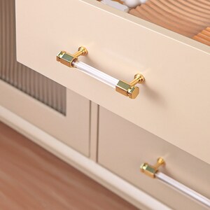 Puede incluir: Dos tiradores de armario dorados con manijas de acrílico transparente. Los tiradores están montados en una puerta de armario blanca.