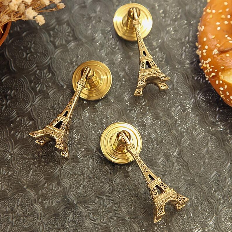 Peut inclure: Trois boutons de meuble dor&eacute;s repr&eacute;sentant la Tour Eiffel. Les boutons ont une base circulaire et une miniature d&eacute;taill&eacute;e de la Tour Eiffel qui s'&eacute;tend vers l'ext&eacute;rieur. Les boutons sont dispos&eacute;s sur une surface grise textur&eacute;e.