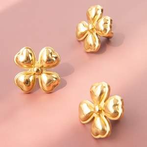 May include: Three gold-colored flower-shaped earrings with a raised center. The earrings are on a pink background.