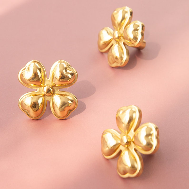 May include: Three gold-colored flower-shaped earrings with a raised center. The earrings are on a pink background.