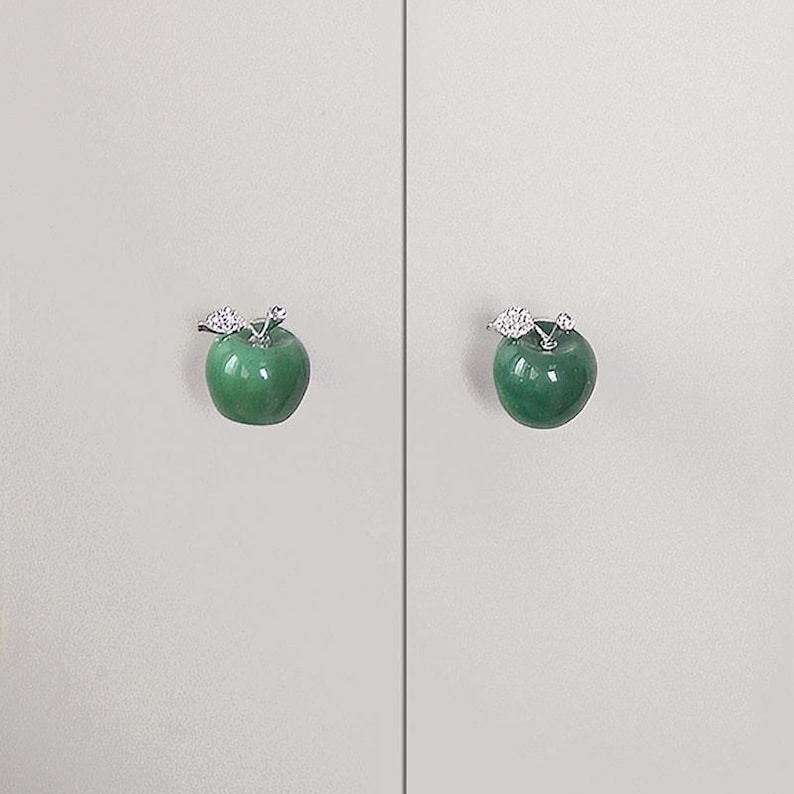 K&ouml;nnte beinhalten: Zwei gr&uuml;ne, apfelf&ouml;rmige Schrankkn&ouml;pfe mit silberfarbenen Blattakzenten. Die Kn&ouml;pfe sind auf einer hellgrauen Oberfl&auml;che montiert und bieten eine dekorative und funktionale Hardware-L&ouml;sung f&uuml;r M&ouml;bel.