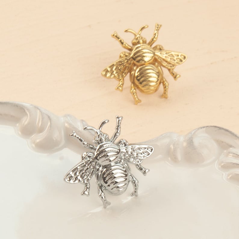 Peut inclure: Deux broches en forme d'abeille, l'une dor&eacute;e et l'autre argent&eacute;e, avec des ailes d&eacute;taill&eacute;es et un corps textur&eacute;. Les broches sont sur une surface blanche.