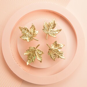 Puede incluir: Cuatro tiradores de caj&oacute;n en forma de hoja de arce de color dorado sobre un fondo rosa.