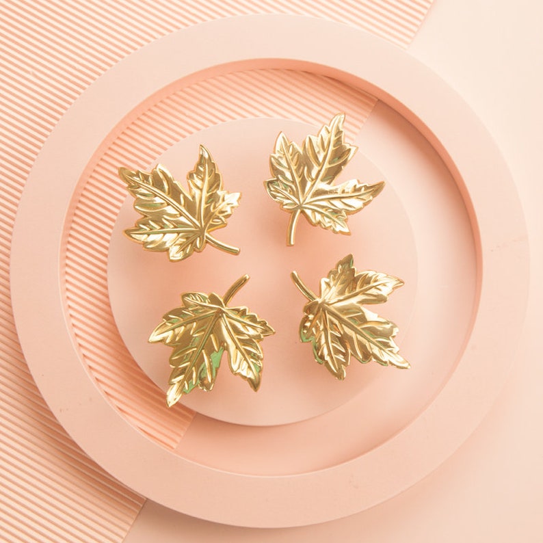 Puede incluir: Cuatro tiradores de caj&oacute;n en forma de hoja de arce de color dorado sobre un fondo rosa.