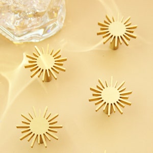 May include: Four gold-toned sunburst cabinet knobs with a circular base. The knobs are arranged in a semi-circle on a light beige surface.