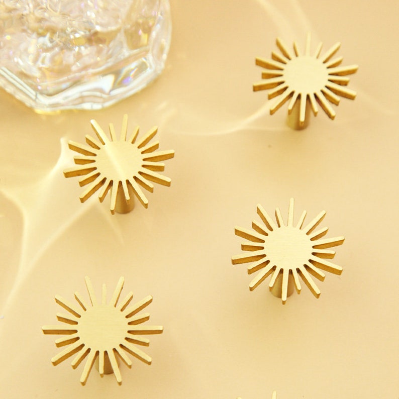 May include: Four gold-toned sunburst cabinet knobs with a circular base. The knobs are arranged in a semi-circle on a light beige surface.