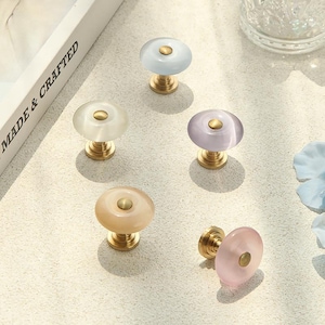 May include: Five round cabinet knobs with smooth, translucent, pastel-coloured tops in light blue, white, lavender, peach, and pink. Each knob features a central circular brass-toned metal accent and a matching brass-toned base for mounting.
