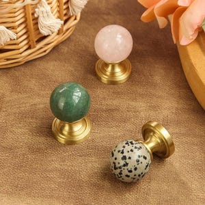 Può includere: Una collezione di pomelli decorativi per mobili. I pomelli presentano top sferici in pietra verde, rosa e maculata, montati su basi in ottone. Questi pomelli sono progettati per mobili e decorazioni per la casa.