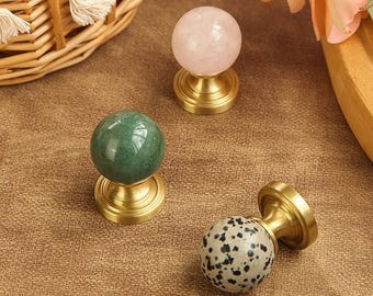 May include: A collection of decorative cabinet knobs. The knobs feature spherical stone tops in green, pink, and speckled patterns, mounted on brass bases. The knobs are designed for furniture and home decor.