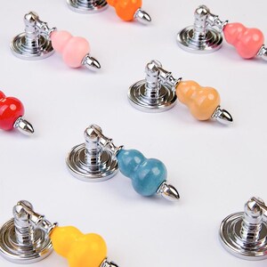 May include: A selection of colourful gourd-shaped cabinet knobs. Each knob has a silver-toned base and a decorative gourd in various colours, including pink, orange, yellow, blue, and red. The knobs are arranged on a white surface.