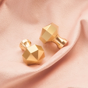 以下が含まれることがあります： ファセットデザインの金色の幾何学的なキャビネットノブ2個。ノブはピンクの布地の背景の上にあります。