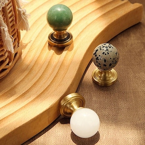 Puede incluir: Tres pomos decorativos para armarios con bases de latón y tapas esféricas. Un pomo tiene una tapa de piedra verde, otro una tapa de piedra gris moteada y el tercero una tapa blanca opaca. Los pomos se exhiben sobre una superficie de madera.
