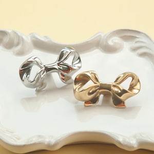 May include: Two decorative bow-shaped drawer pulls, one silver and one gold, on a white ceramic tray.