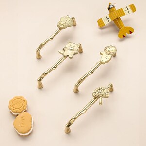 May include: Gold-coloured cabinet pulls featuring animal designs, such as a lion, bear, and turtle. The pulls have a branch-like handle. A yellow toy aeroplane and two sandwich biscuits are also in the image.