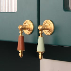 May include: Close-up of two cabinet pulls. Each pull has a gold-colored circular backplate and a decorative handle. One handle is brown, the other is light green. The pulls are mounted on a teal-colored cabinet door.