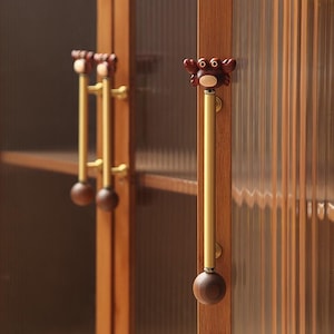 Peut inclure: Une poignée de placard composée d'une barre verticale en laiton avec une sphère en bois brun foncé à la base et un ornement en bois brun foncé en forme de crabe au sommet, fixée sur une porte de placard en bois avec des panneaux de verre.
