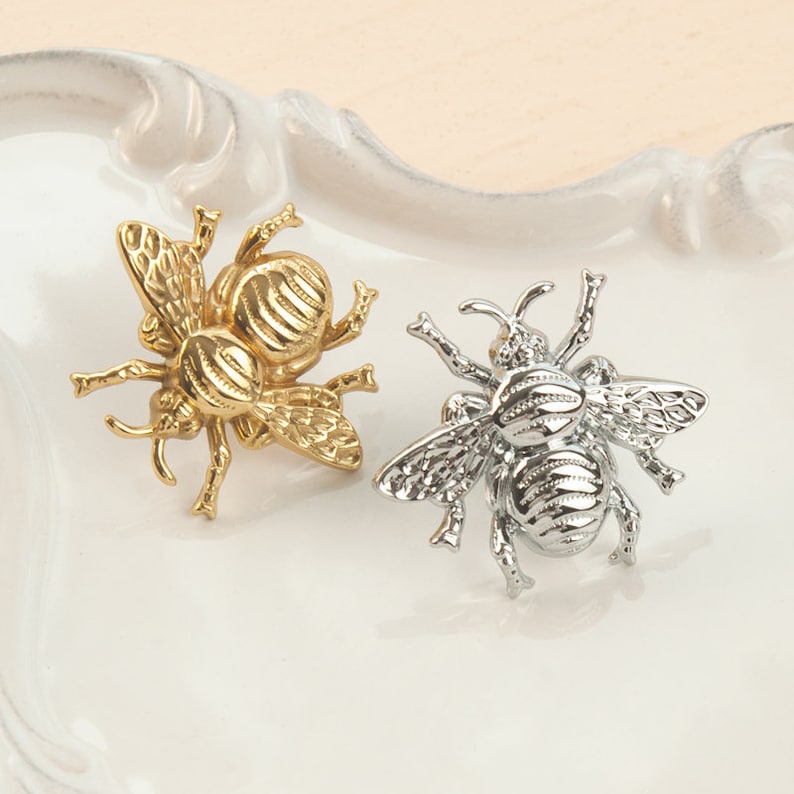 Peut inclure: Deux anneaux de serviette en m&eacute;tal en forme d'abeille, l'un dor&eacute; et l'autre argent&eacute;, sur un fond blanc.