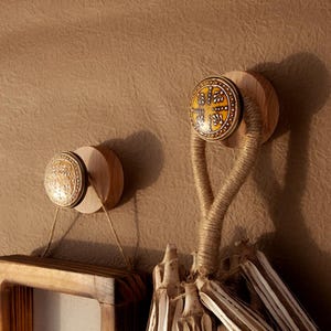 May include: Two decorative wall hooks with wooden bases and ceramic knobs. One knob features a yellow and brown cross design, while the other has a circular pattern. Natural twine and wood elements create a rustic aesthetic.