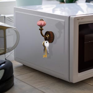 Könnte beinhalten: Ein dekorativer Schlüsselhaken mit einem rosa Keramikknauf und einem Messinghaken, montiert auf einem Holzsockel. Mehrere Schlüssel hängen an dem Haken, befestigt an einer weißen Mikrowelle.