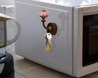 Puede incluir: Un colgador de llaves decorativo con un pomo de cerámica rosa y un gancho de latón, montado sobre una base de madera. Varias llaves cuelgan del gancho, fijadas a un microondas blanco.