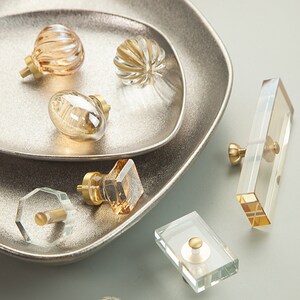 May include: Assortment of decorative cabinet knobs with glass and metal accents. The knobs are in various shapes and sizes, including round, oval, square, and rectangular. The knobs are in shades of gold and clear glass.
