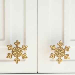 Op de afbeelding: Twee gouden sneeuwvlok-kastknoppen op een witte kastdeur.