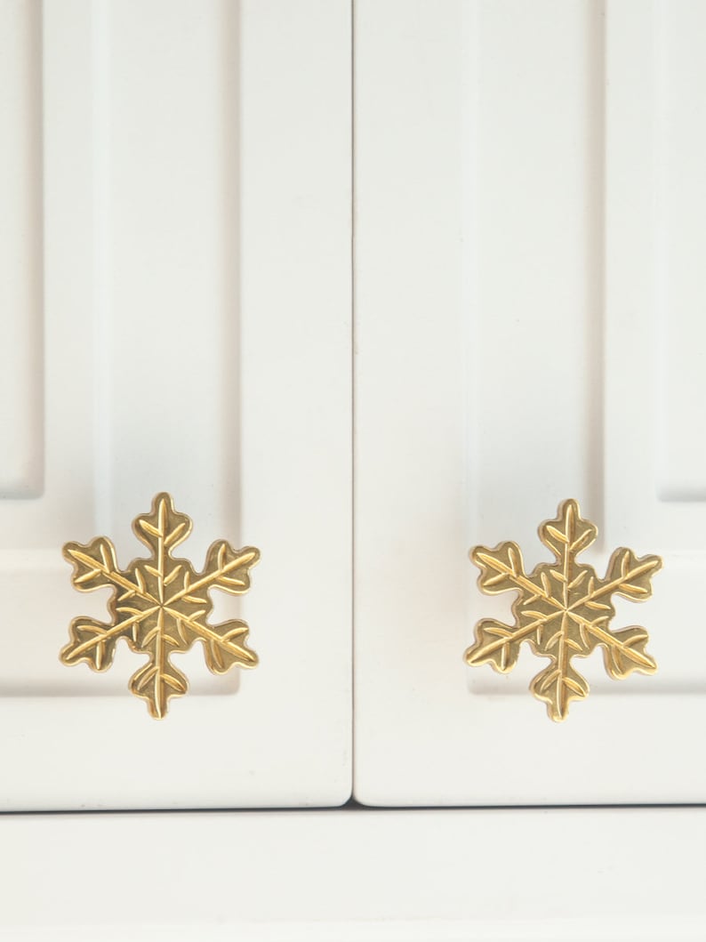 Op de afbeelding: Twee gouden sneeuwvlok-kastknoppen op een witte kastdeur.