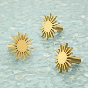 May include: Three gold-toned sunburst-shaped cabinet knobs with a circular center and a raised, textured design. The knobs are arranged in a triangular pattern on a light blue textured surface.